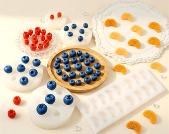 Può includere: Una collezione di stampi in silicone per creare forme di frutta in miniatura, inclusi stampi rotondi per mirtilli e lamponi, e stampi rettangolari per spicchi d'arancia, accompagnati da repliche di frutta blu e arancione.