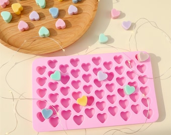 May include: A rectangular pink silicone mold featuring multiple heart-shaped cavities, accompanied by a wooden plate holding various small, pastel-colored heart-shaped pieces in shades of blue, yellow, pink, and green, with thin fairy lights.