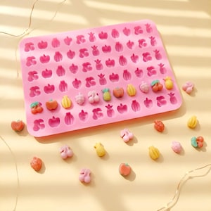 Peut inclure: Un moule en silicone rose rempli de diverses formes de fruits, dont des pommes, des poires et des cerises. Plusieurs bonbons colorés en forme de fruits sont éparpillés autour du moule. Le moule est conçu pour faire des friandises en forme de fruits.