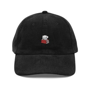 May include: A black corduroy baseball cap featuring a small embroidered design on the front center. The embroidery depicts a white puppy resting its head on a stack of three red books with white pages. The cap has a curved brim and a button top.