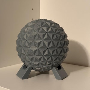 May include: A gray geometric sphere with a triangular pattern, resting on a matching gray stand. The sphere is approximately 6 inches in diameter. The design is modern and minimalist, suitable for home decor.