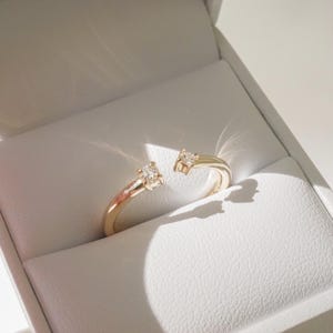 May include: Open-ended gold ring with two clear, square-cut gemstones. The ring is displayed inside a white jewelry box with a textured interior. The ring's design allows for size adjustment.