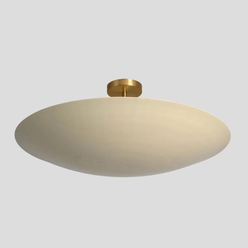 May include: Cream-colored, oval-shaped ceiling light fixture with a brass-colored base. The light has a smooth, rounded design, and is mounted on a white ceiling. The fixture is a modern design.