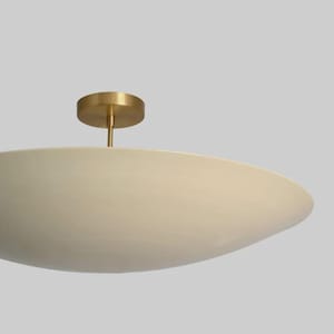 May include: A modern, cream-colored ceiling light fixture with a brass-colored base and stem. The light has a wide, shallow bowl shape, and is designed to be mounted flush against the ceiling. The fixture is a simple, elegant design.