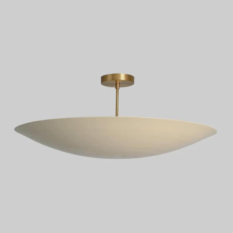 May include: A modern, cream-colored ceiling light fixture with a wide, shallow, bowl-shaped shade. The fixture has a brass-colored stem and a circular ceiling mount. The design is minimalist and elegant, suitable for various interior styles.