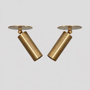 May include: Two brass-colored, cylindrical spotlights with a circular base and a ball joint. The spotlights are angled and appear to be made of metal. The background is plain white.