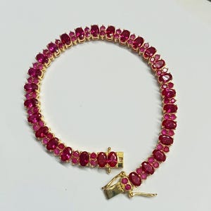 May include: A gold-toned bracelet featuring a series of oval-cut, vibrant red gemstones. The gemstones are closely set, creating a continuous line around the wrist. The clasp is also gold-toned, with a secure closure.