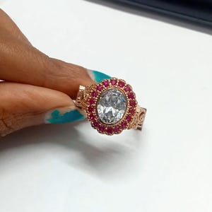 May include: Rose gold ring featuring a large, oval-cut, clear gemstone surrounded by a halo of small, red gemstones. The ring has intricate filigree detailing on the band and around the halo.