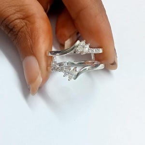 Peut inclure: Une protection de bague en argent avec un design incurvé, ornée de pierres précieuses transparentes. La protection de bague a une tige fendue et est conçue pour être portée avec une autre bague. Les pierres sont serties en pavé et en marquise.