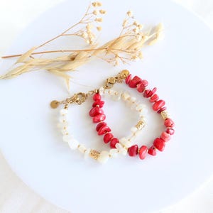 Peut inclure: Deux bracelets de perles. L'un présente des pierres rouges ressemblant à du corail, l'autre des pierres blanches. Les deux bracelets ont des chaînes et des fermoirs dorés. Les bracelets sont présentés sur une surface blanche avec des accents floraux séchés.