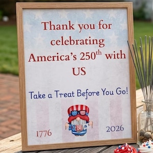 Pode incluir: Uma placa emoldurada em madeira com o texto "Thank you for celebrating America's 250th with US" e "Take a Treat Before You Go!" é exibida em uma mesa de piquenique de madeira. Cupcakes vermelhos, brancos e azuis estão em um prato. Também são visíveis sparkles e bebidas.