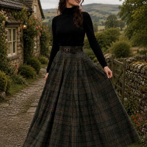 May include: A black long-sleeve turtleneck sweater paired with a floor-length, pleated, dark green and brown plaid wool skirt. The skirt features a high waist cinched with a dark brown leather belt that has a decorative metal buckle.