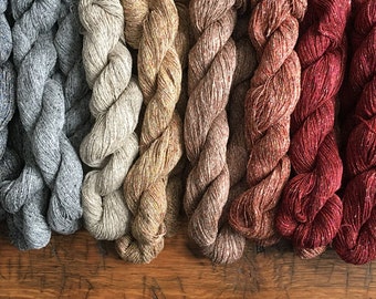 May include: A collection of ten skeins of yarn in various shades of blue, brown, red, and purple. The yarn is a blend of linen and wool and has a speckled appearance.