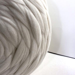 May include: Close-up of a large, off-white ball of thick yarn. The yarn has a soft, textured appearance with visible strands. The background is a plain white surface, providing contrast to the yarn's texture. Ideal for crafting projects.