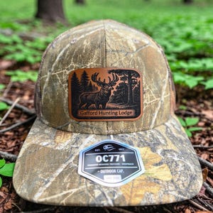 May include: A camouflage print trucker cap featuring a rectangular brown leather patch with a black engraved graphic of a deer in a forest and the text Gafford Hunting Lodge. A hexagonal sticker on the brim displays the text OC771 and Outdoor Cap.