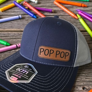 May include: Navy blue trucker hat with a light gray mesh back and a brown leather patch that reads "POP POP". The hat has a curved brim and a snapback closure. A sticker on the hat reads "OC771". Colorful crayons are scattered around the hat.