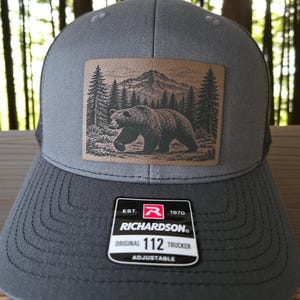 May include: A grey trucker hat featuring a rectangular tan patch with a black graphic of a bear walking through a forest with a mountain in the background. A black and white sticker on the brim reads EST. 1970 RICHARDSON ORIGINAL 112 TRUCKER ADJUSTABLE.