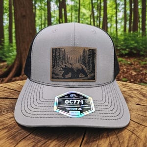 May include: A gray trucker hat with a black mesh back and a curved brim. The hat features a rectangular patch with a bear walking through a forest. The patch is brown and tan. The hat has the text "OC771" and "Outdoor Cap".
