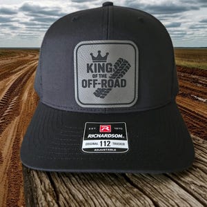 May include: A charcoal gray trucker hat featuring a square patch with a crown icon, tire tread graphic, and the text KING OF THE OFF-ROAD. A black and white sticker on the brim displays the Richardson 112 Original Adjustable branding.
