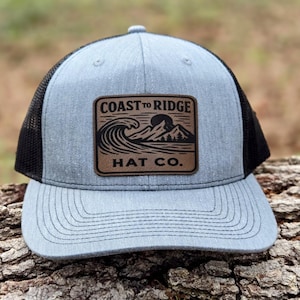 May include: A light blue trucker hat with a black mesh back. The hat features a brown leather patch with the words "COAST TO RIDGE HAT CO." and a graphic of a wave, mountains, and a sun.