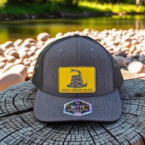 May include: A gray and black trucker hat with a yellow patch featuring a coiled snake and the text "DON'T TREAD ON ME." The hat has a mesh back and a curved brim. The hat also has a rectangular tag with the text "OC771".
