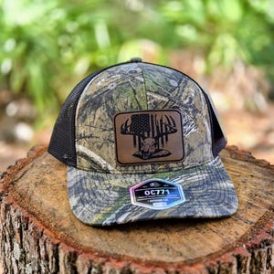 May include: Camouflage baseball cap with a black mesh back. The front features a leather patch with a deer skull and an American flag design. The brim has a camouflage pattern, and the cap includes the text "OC771".