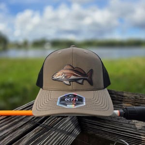 May include: A tan and black baseball cap featuring a detailed graphic of a fish on the front panel. The brim displays a hexagonal sticker with the text OC771. The cap has a structured crown and a curved visor with visible stitching along the edge.