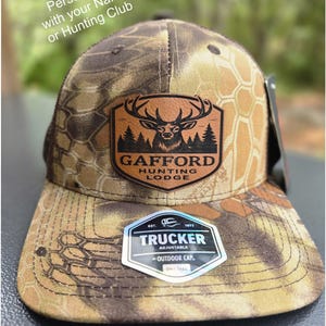 May include: Camouflage trucker hat with a brown leather patch featuring a deer head and the text "GAFFORD HUNTING LODGE." The hat has a mesh back and a curved brim. The tag reads "TRUCKER ADJUSTABLE OUTDOOR CAP."