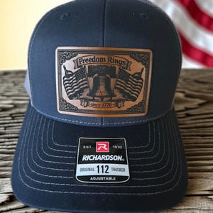 May include: A navy blue trucker hat featuring a rectangular brown leather patch with an engraved Liberty Bell, two American flags, and the text Freedom Rings since 1776. A black sticker on the brim displays the Richardson 112 Original Trucker logo.