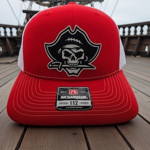 May include: Red and white trucker hat with a pirate skull logo. The hat features a red front panel and brim, a white mesh back, and a black and white pirate skull design on the front. The hat has a curved brim and a snapback closure.