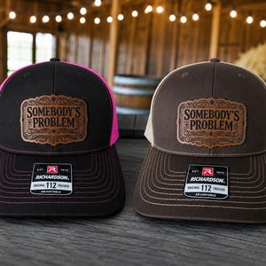 May include: Two adjustable trucker hats featuring a leather patch with the text SOMEBODY'S PROBLEM. The left hat is black with bright pink mesh, and the right hat is brown with tan mesh. Both hats display a Richardson 112 brand sticker on the brim.