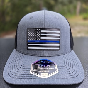 May include: Gray trucker hat with a black mesh back and a flat brim. The front features a patch of the American flag with a blue line. A holographic sticker on the brim reads "OC771 PREMIUM MODERN TRUCKER - SNAPBACK OUTDOOR CAP ONE SIZE."