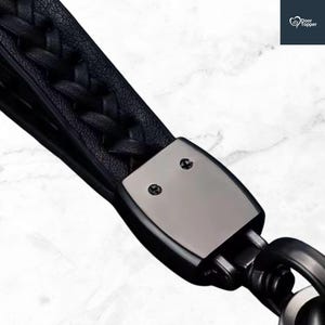 May include: Black leather keychain with a braided strap and a gunmetal-coloured metal clasp. The keychain has a sturdy metal ring for attaching keys. The design is sleek and modern, suitable for everyday use.