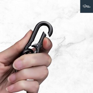 May include: A black metal carabiner clip, held in a hand, against a white marble background. The clip is open, showing its spring-loaded mechanism. The Door Topper logo is in the upper right corner.