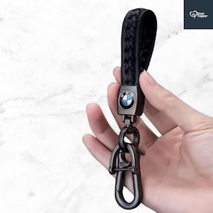 May include: Black leather braided keychain with a metal clasp and a BMW logo. The keychain is held in a hand against a white marble background. The BMW logo is a blue and white circle.