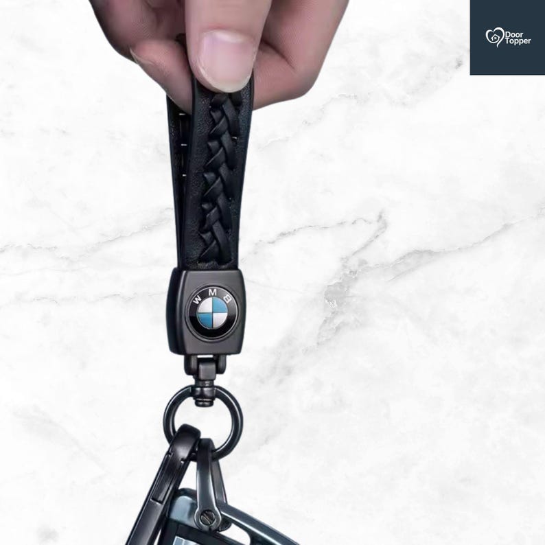 May include: Black leather braided keychain with a BMW logo. The keychain has a metal clasp and a split ring for attaching keys. The BMW logo is a circular emblem with blue and white segments. The keychain is held by a hand.