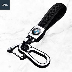 May include: Black leather braided keychain with a silver clasp and a BMW logo. The keychain has a black leather strap and a metal clasp for attaching keys. The BMW logo is in the centre of the metal plate.