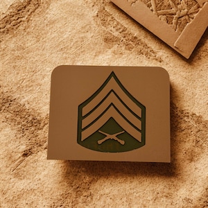 Könnte beinhalten: Ein rechteckiges, hellbraunes militärisches Rangabzeichen mit drei Winkeln und gekreuzten Gewehren. Das Innere ist dunkelgrün. Das Design ist vor einem strukturierten, hellbraunen Hintergrund platziert.