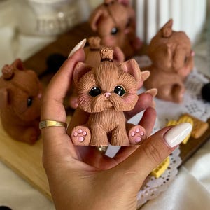 Puede incluir: Una colección de figuras de gatos marrones con ojos grandes y detalles rosas. Las figuras se exhiben en una tabla de madera con elementos decorativos. Las figuras de gatos tienen una apariencia linda y de dibujos animados.