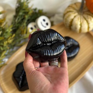 Puede incluir: Objetos decorativos negros con forma de labios sobre una bandeja de madera. Los labios brillantes tienen temática de Halloween, con calabazas y otras decoraciones otoñales de fondo. Los labios son de un negro oscuro y brillante.