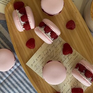 Könnte beinhalten: Rosa Macarons, gefüllt mit weißer Creme und frischen Himbeeren, sind auf einem Holztablett angeordnet. Mehrere ganze Himbeeren sind um die Macarons verstreut. Die Szene spielt sich auf einer blau-weiß gestreiften Oberfläche ab.