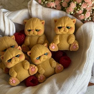 Könnte beinhalten: Fünf gelbe katzenförmige Figuren mit grimmigen Ausdrücken, rosa Wangen und blauen Augen liegen in einem mit weißem Stoff ausgekleideten Korb mit roten Beeren. Die Figuren haben detaillierte Pfoten und sind von rosa Blumen umgeben.