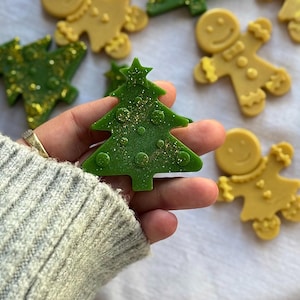 May include: A close-up of a green Christmas tree-shaped wax melt with gold glitter, held in a hand. Other wax melts in the shape of Christmas trees and gingerbread people are scattered in the background.