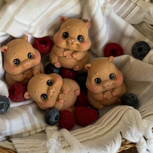 Könnte beinhalten: Eine Ansammlung brauner Hamsterfiguren mit großen schwarzen Augen und rosigen Wangen, eingebettet zwischen frischen Himbeeren und Blaubeeren in einem Weidenkorb, der mit einem cremefarbenen Tuch ausgekleidet ist.