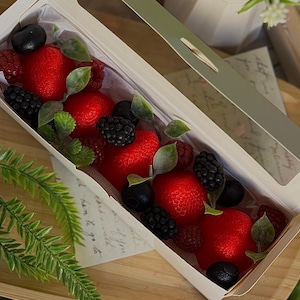 Puede incluir: Una caja rectangular con tapa transparente, llena de fresas rojas, moras, frambuesas y arándanos. Hojas verdes se intercalan entre la fruta. La caja está sobre una superficie de madera.