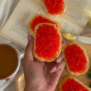 Puede incluir: Una mano sosteniendo una rebanada de pan cubierta con caviar rojo brillante. Varias otras rebanadas de pan cubiertas con caviar están dispuestas en una tabla de madera. También se ven una taza de té, un libro y un plato pequeño con mantequilla.