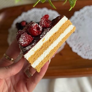 Puede incluir: Una porción de pastel en capas con capas alternas de color beige y blanco, cubierta con frambuesas rojas frescas y un glaseado de chocolate negro. El pastel está espolvoreado con azúcar glas y está sobre una bandeja de madera.