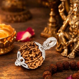 May include: A pendant featuring a textured, brown seed with a silver filigree setting. A lit candle in a gold holder, a small statue, and prayer beads are in the background. The scene is set on a wooden surface.