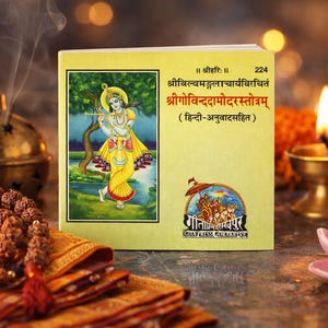 May include: A book with a yellow cover featuring an illustration of a figure playing a flute, with text in a foreign language. Incense smoke rises near a decorative brass incense burner and a strand of prayer beads. A lit candle and a pink lotus flower are also visible.