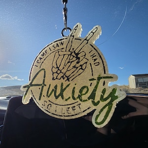 May include: A round, cream-colored car air freshener with the words "I came I saw I had Anxiety so I left" in green and black lettering. The design includes a skeleton hand making a peace sign.